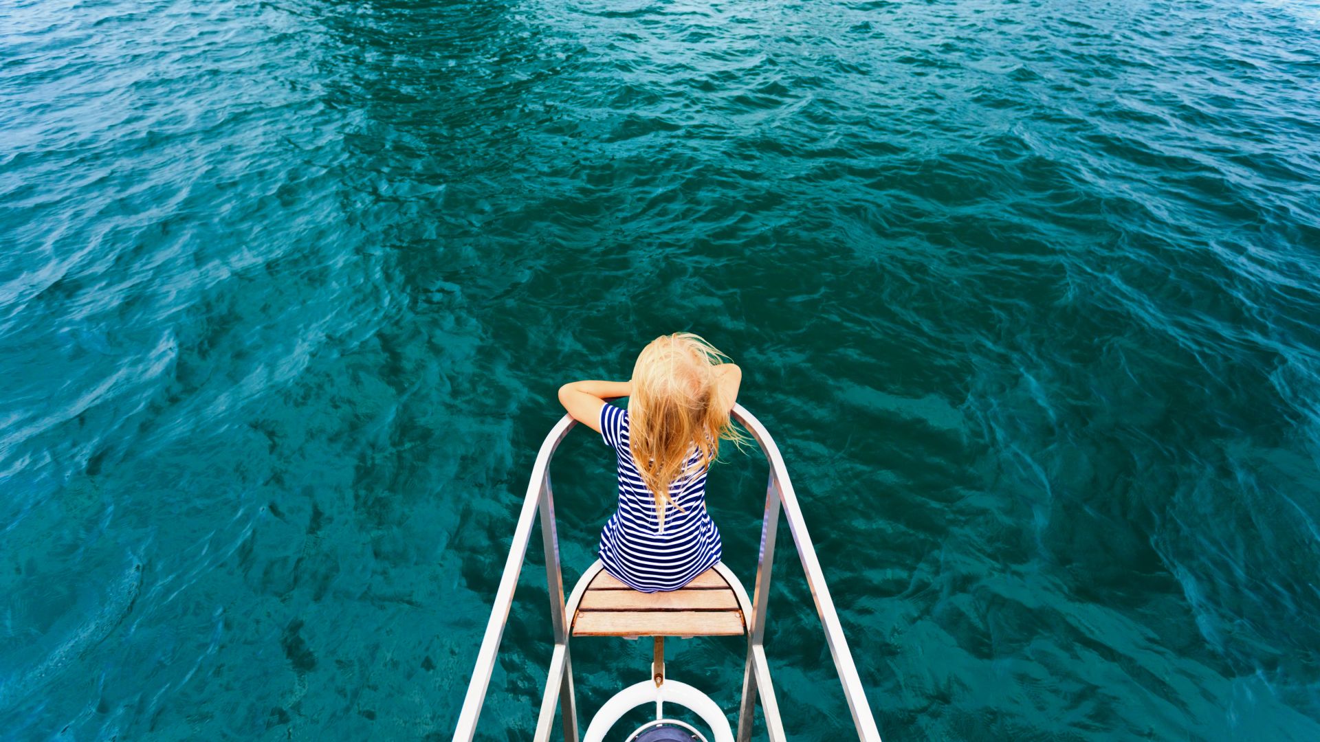A fascinating image shows a young girl on a tranquil sailing boat, sitting in the bow in a white and blue striped swimsuit. The focus of the image is solely on her, lost in the midst of the deep blue sea, evoking a sense of calm and solitude.